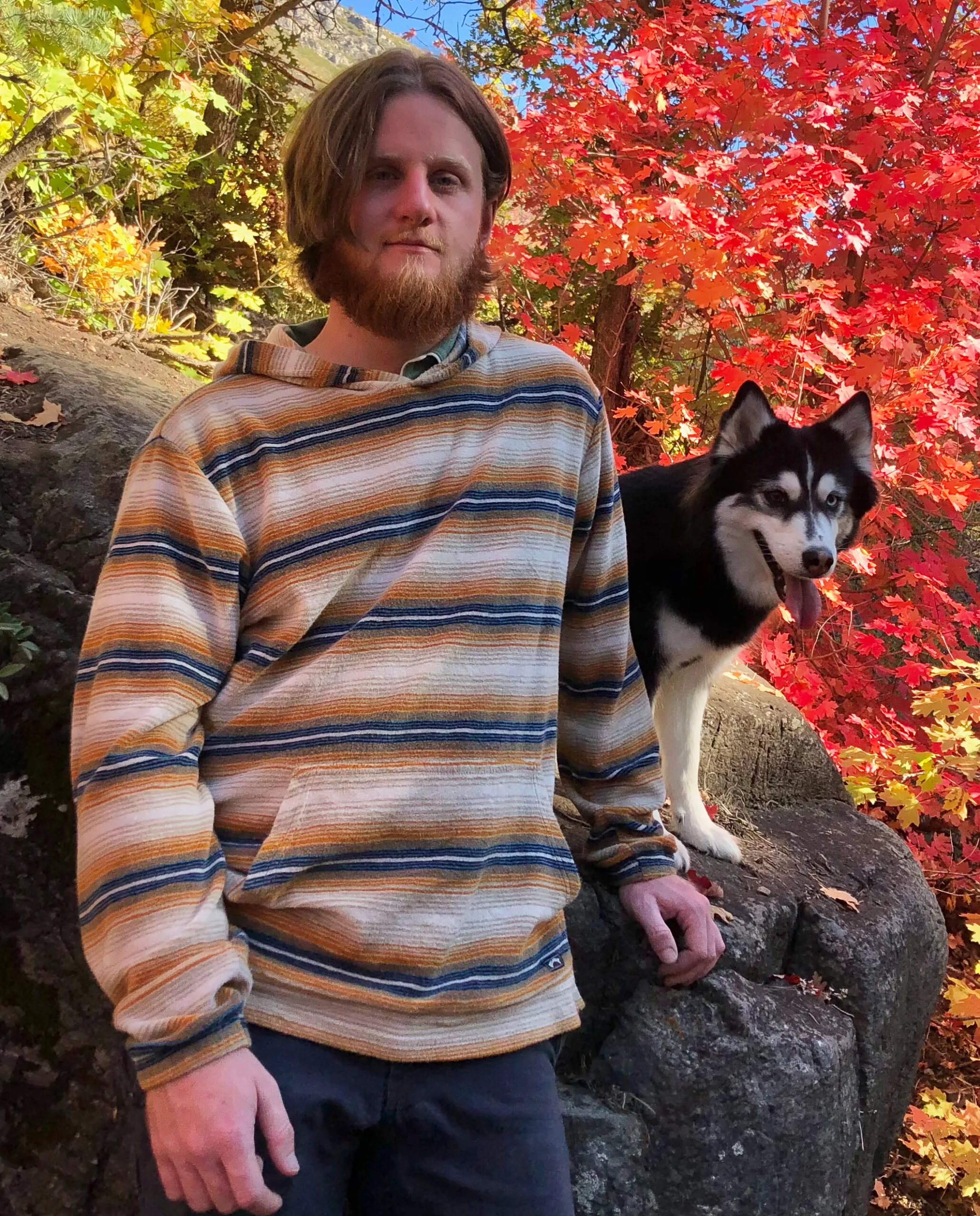 Dillan Knudsen A dog trainer standing outdoors beside a Siberian Husky, surrounded by vibrant autumn foliage and natural rock formations, representing professional canine training, obedience expertise, and an approachable, nature-focused training philosophy.